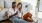 a man and woman with take out, sit against a couch on their living room floor and watch at their cell phone